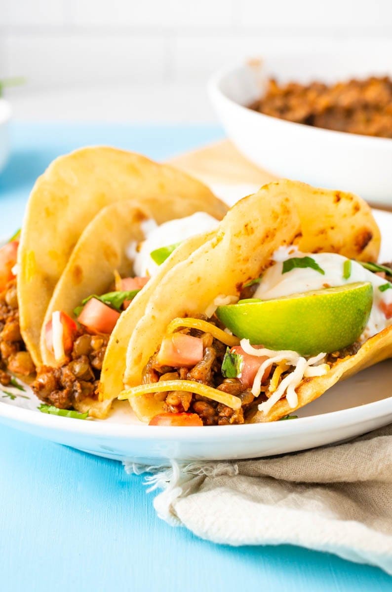 Close-up of three lentil tacos garnished with lime wedges, sour cream, cheese, and diced tomatoes on a white plate.