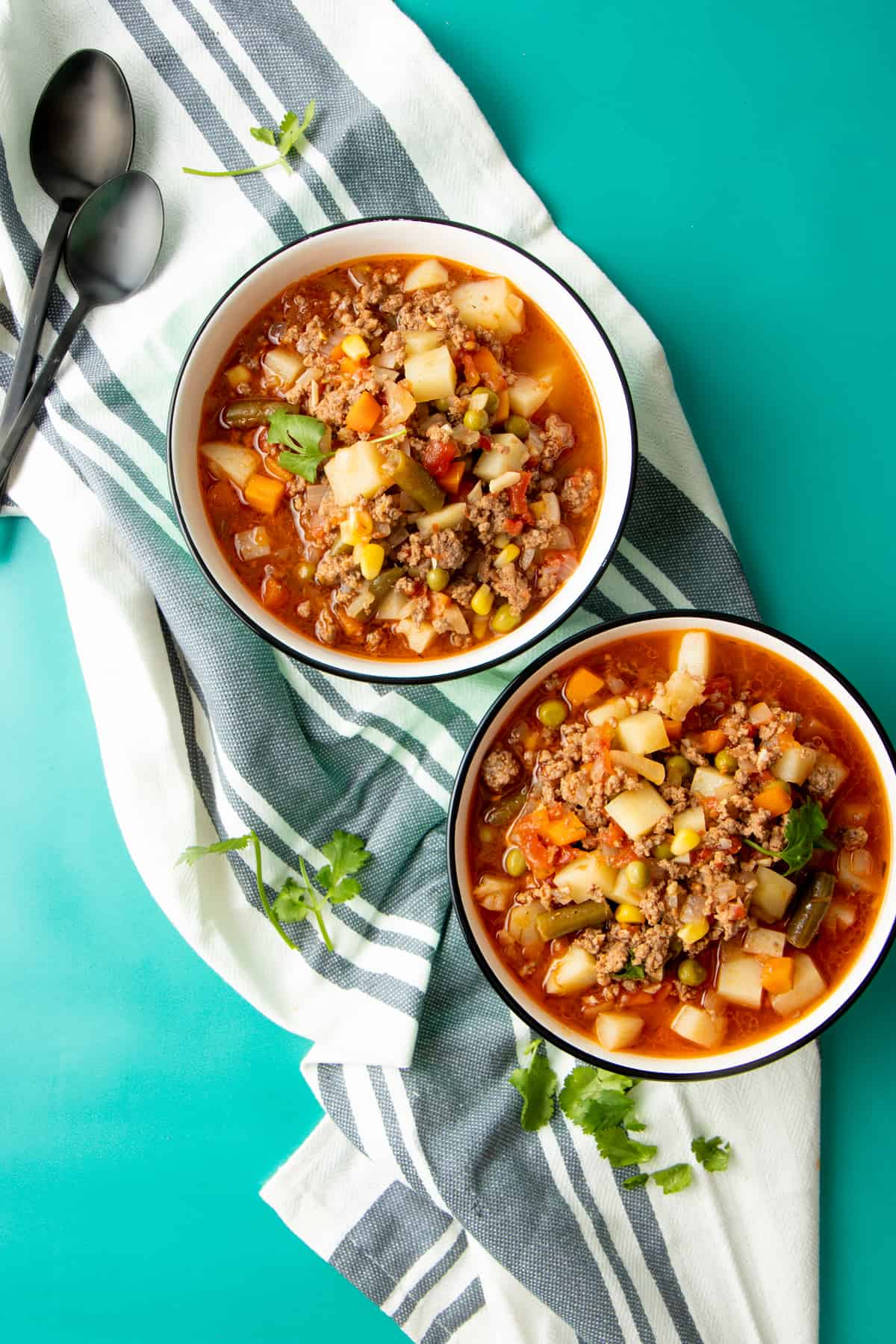 Two bowls of vegetable beef soup sit on a dish towel, with spoons nearby.