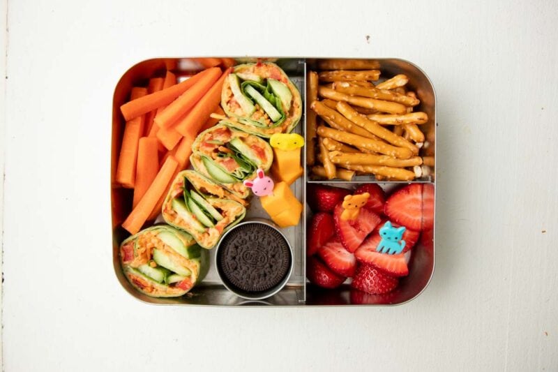 Overhead of bento-style school lunch packed with veggie pinwheels, strawberries, carrots, and pretzels.