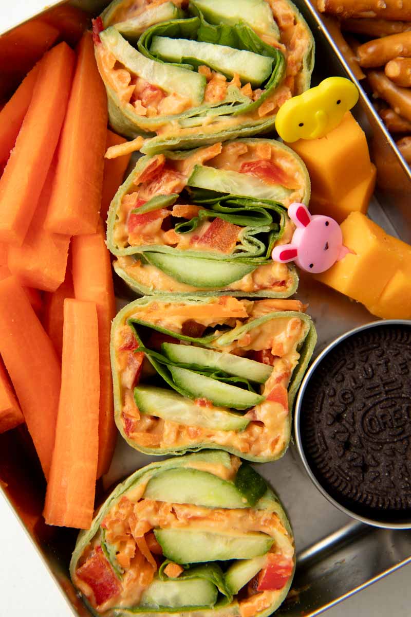 Close view of four veggie pinwheels in the large compartment of a lunchbox with carrot sticks, cheese cubes, and an oreo cookie alongside.