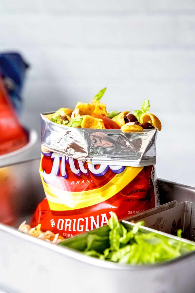 A corn chip snack bag stands up in a lunch box with the makings of a walking taco visible from the top of the bag, including corn chips, lettuce, tomatoes, black beans, and seasoned beef.