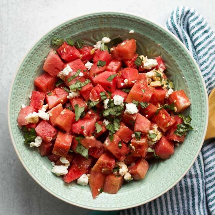 A wooden spoon rests on a striped linen napkin beside a decorative bowl filled with watermelon basil salad.