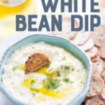 White bean dip drizzled with olive oil is in a light blue bowl, surrounded by crackers. A text overlay reads "Roasted Garlic White Bean Dip."